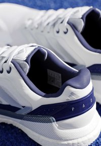 Chaussures de sport blanches et bleu marine avec lacets, placées sur une surface texturée bleue, montrant les détails du talon et du côté.