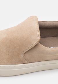 Beige Wildleder Slipper mit runder Zehenpartie, gesteppten Akzenten und einer leichten Gummisohle. Das Innenfutter besteht aus einem weichen braunen Stoff.