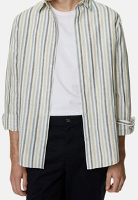 Camicia a righe in tessuto leggero, con righe blu, grigie e crema. Maniche lunghe arrotolate ai polsi, canottiera bianca visibile.