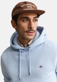 Homme portant un sweat à capuche bleu clair avec le logo GANT et une casquette marron avec un texte blanc, regardant légèrement vers la droite sur un fond uni.