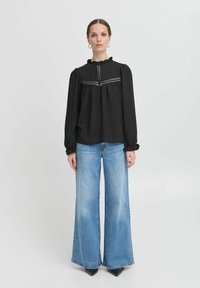 Top nero a maniche lunghe con colletto arricciato e cuciture decorative. Abbinato a jeans blu a gamba larga e scarpe nere a punta.