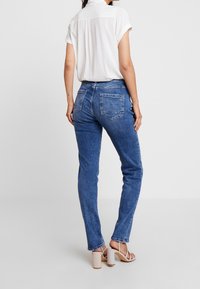 Vaqueros de mezclilla azul con un corte ajustado, cinco bolsillos y detalles de costura. Combinados con una blusa blanca de manga corta y sandalias de tacón cuadrado beige.