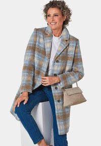 Karierter Wollmantel in Blau-, Braun- und Cremetönen; mit großen Knöpfen und einem Reverskragen. Kombiniert mit Jeans und einer beigefarbenen Handtasche.
