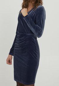 Marineblaues Samtkleid mit langen Ärmeln, V-Ausschnitt und gerafftem Detail an der Taille. Glatte Textur, knielanger Schnitt.