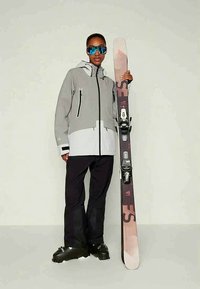 Άτομο με γκρι και λευκή σακάκι σκι, μαύρα παντελόνια και μπότες σκι, κρατώντας ένα μόνο όρθιο σκι με σταθεροποιήσεις, φορώντας ανακλαστικά γυαλιά σκι.