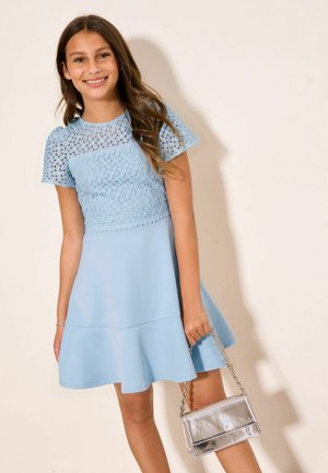 Smiling girl in blue lace top dress holding a small silver purse against a plain light background.