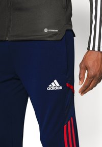 Černá bunda Aeroready, námořnické kalhoty s červenými akcenty, logo Adidas, hladká textura, přiléhavý design, viditelné detaily postranních kapes.