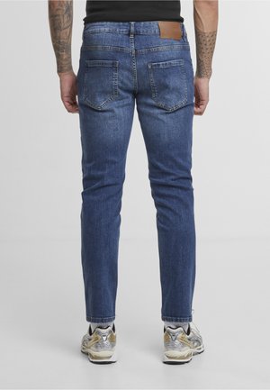 Mann trägt blaue Slim-Fit-Jeans und weiße Sneakers, steht mit dem Rücken zum Betrachter, tätowierte Arme vor einfarbigem Hintergrund sichtbar.