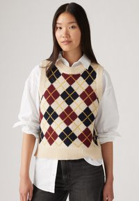 Strikket genser med argyle mønster i kremfarge, med røde, marineblå og gule diamanter, med rund hals og ribbestrikket kant, lagd over en hvit skjorte.
