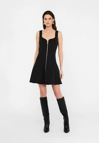 Czarna sukienka bez rękawów z dopasowanym górnym partem i rozkloszowanym dołem; posiada zamek błyskawiczny z przodu i gładką fakturę tkaniny. Czarna, sięgająca do kolan obuwie.