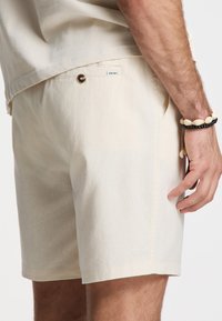 Light beige shorts made of soft fabric, featuring a button detail, side pockets, and a relaxed fit, with a smooth texture.