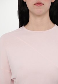 Light pink long-sleeve top with a round neckline, featuring diagonal seam details and a smooth, soft texture. No patterns or embellishments.