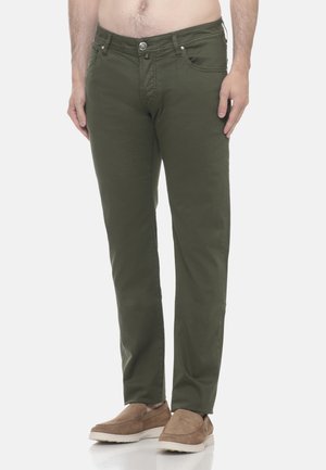 Uomo che indossa pantaloni verde oliva a gamba dritta e scarpe marroni slip-on, in piedi su uno sfondo bianco uniforme.