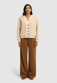 Cardigan in pile crema con scollatura a V, cinque bottoni marroni e polsini elasticizzati, abbinato a pantaloni larghi marroni e scarpe slip-on con motivo.