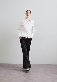 Camicia bianca con bottoni e maniche lunghe, abbinata a pantaloni neri in satin e scarpe col tacco nere, su uno sfondo neutro.