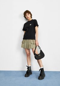 T-shirt en coton noir avec une poche poitrine, jupe plissée verte olive avec poches cargo, bottes de combat noires et un petit sac à main noir.