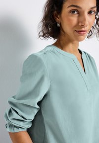 Femme aux cheveux bouclés et foncés, portant un blouse légère à manches longues de couleur bleu clair et des boucles d'oreilles argentées, se tenant devant un mur blanc uni.