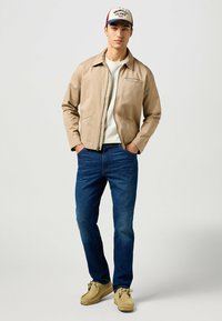 Lys beige zip-up jakke med spids krave, hvid t-shirt under, mørkeblå jeans og brune suede loafers. En afslappet cap fuldender looket.