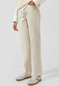 Osoba ubrana w beżowe spodnie ze sznureczkiem, białą koszulę z długim rękawem i srebrne sneakersy z brązowymi podeszwami na jednolitym tle.