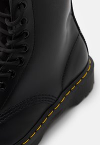 Botte en cuir noir à lacets avec une couture jaune le long de la semelle épaisse en caoutchouc, montrée en gros plan.