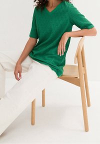 Top in maglia verde a maniche corte e scollo a V con motivo ad occhiello, abbinato a pantaloni dritti color crema, seduto su una sedia di legno.