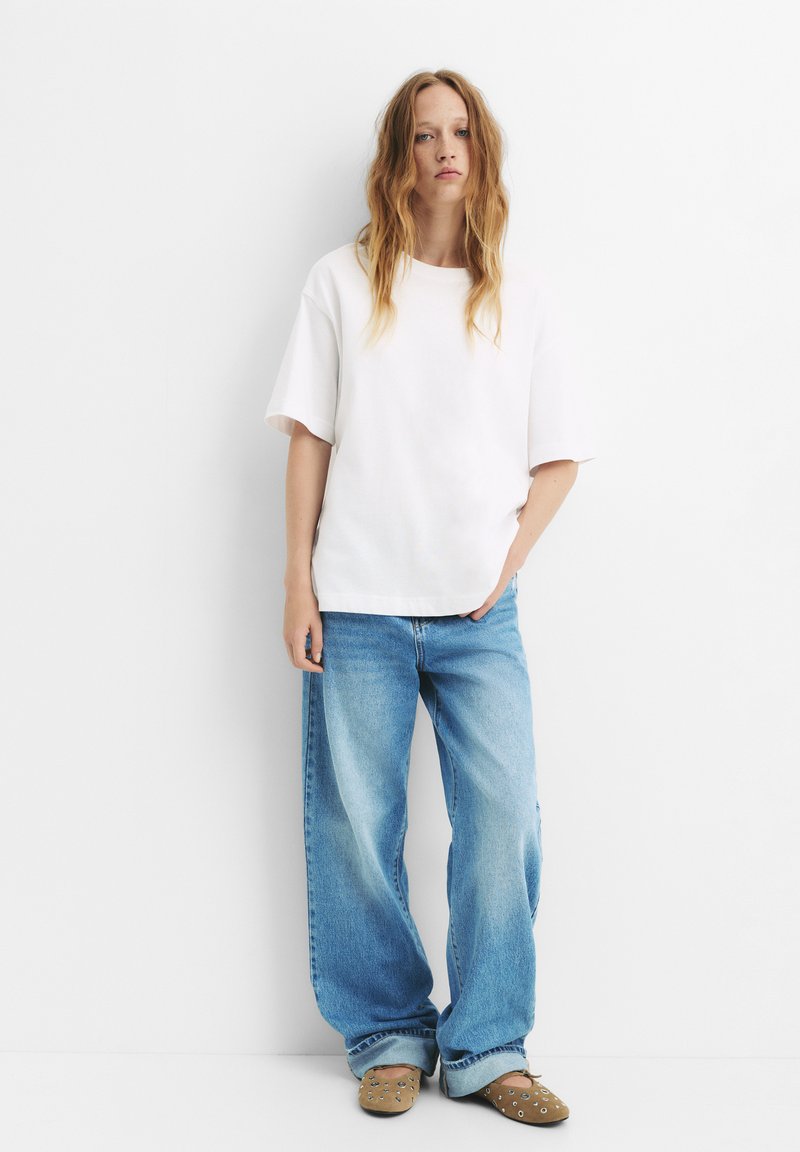 Bela bombažna majica s kratkimi rokavi, v kombinaciji s poledi modrimi oversized denim hlačami in svetlo rjavimi natikači s konicami.