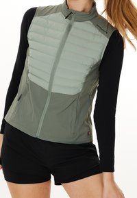 Lehká olivově zelená vesta s prošívanou horní částí, s elastickým zapínáním na zip, bočními kapsami a elegantním designem. V kombinaci s černými šortkami.