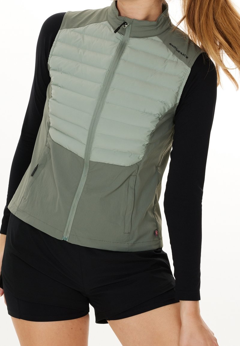 Lehká olivově zelená vesta s prošívanou horní částí, s elastickým zapínáním na zip, bočními kapsami a elegantním designem. V kombinaci s černými šortkami.