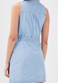 Robe en denim légère sans manches de couleur bleu clair avec une ceinture, vue de dos sur une personne contre un fond blanc uni.