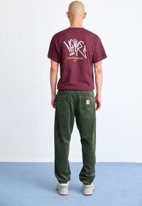 Maroon graphic t-shirt with white and yellow text on back, paired with green corduroy pants and white sneakers with black accents.