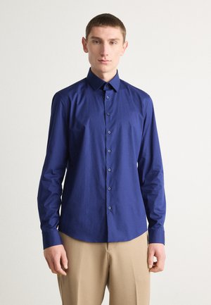 Navy blue long-sleeve shirt with a button-down collar, front buttons, and a smooth texture. Worn with light beige trousers.