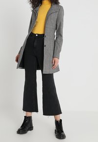 Tjekket grå frakke med en bred krave, parret med en gul turtleneck, sorte cropped jeans og sorte snørebåndsstøvler.
