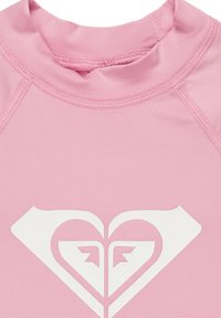 Rosa rash guard med hög krage, korta ärmar och platta sömmar. Har en stor vit hjärtformad logotyp i mitten.