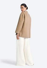 Oversized jas van een wolmix in beige met wijde mouwen, knoopmanchetten en een rugpasdetail, gecombineerd met vloeiende witte broek met wijde pijpen.