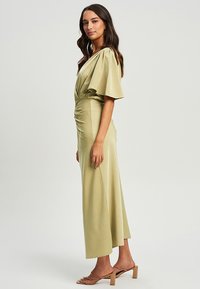 Grünes Satin-Kleid mit kurzen, fließenden Ärmeln, taillierter Taille und seitlichem Raffung, das bis zu einem schwingenden, knöchellangen Saum reicht. Durchsichtige, rückenfreie Sandalen.