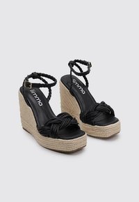 Schwarze geflochtene Wedge-Sandaletten mit einer Plattform im Jutestil, die ein geknotetes Design vorne und verstellbare Knöchelriemen aufweisen.