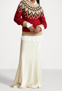 Pull à motifs rouges et beiges avec un col rond, associé à une jupe longue en satin crème et des baskets blanches. Détail en maille texturée.