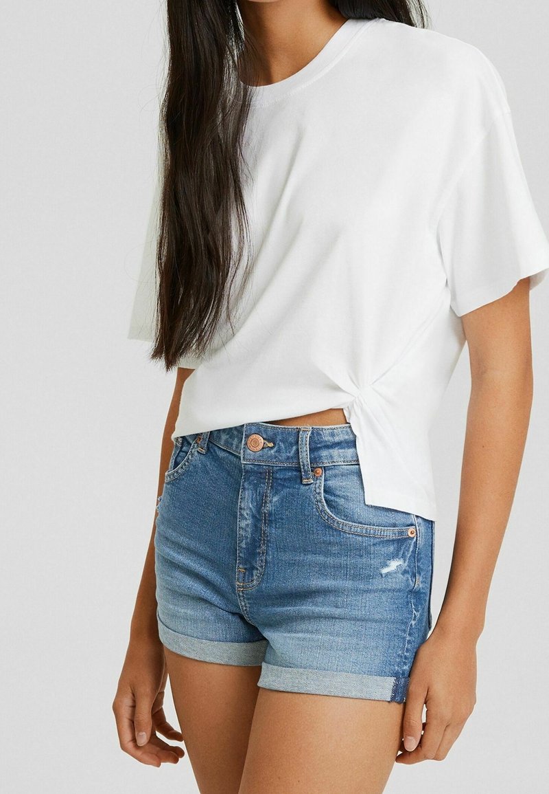 Vit t-shirt med korta ärmar och knutdetalj på sidan. Bärs med blå denimshorts med uppvikta fållar och slitna detaljer.