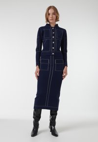 Vestido de punto azul marino con botón en la parte delantera, cuello alto y costuras en contraste. Tiene dos bolsillos frontales y una silueta ajustada.