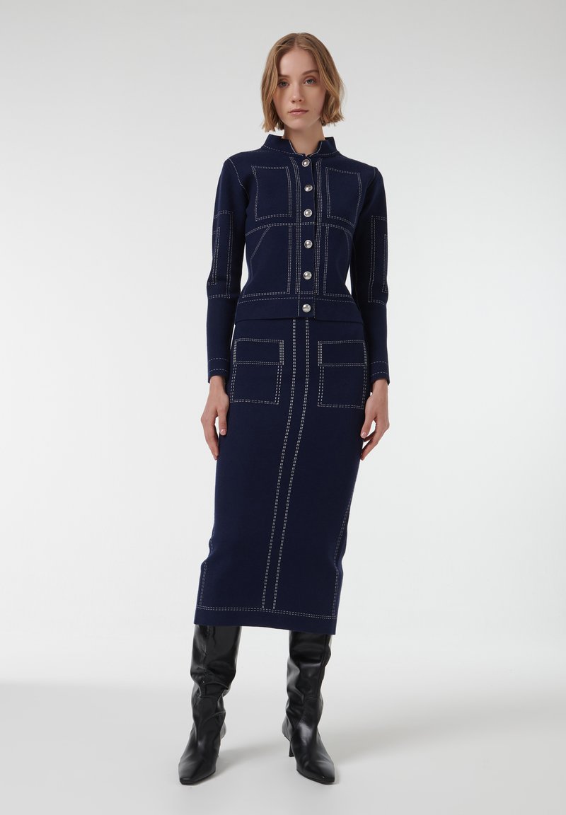 Vestido de punto azul marino con botón en la parte delantera, cuello alto y costuras en contraste. Tiene dos bolsillos frontales y una silueta ajustada.