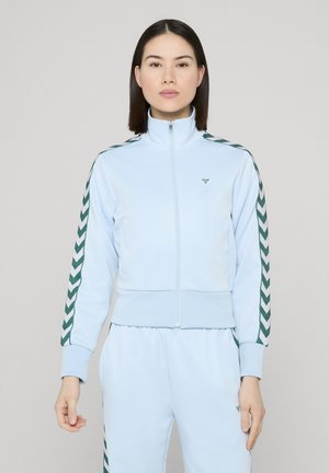 Lyseblå lynlåstrøje lavet af blødt stof med grønne chevronstriber på ærmerne og et lille logo på brystet.