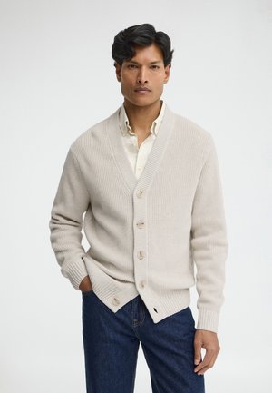 Man die een roomkleurige gebreide cardigan draagt over een wit overhemd met knopen en blauwe spijkerbroek, staand met één hand in de zak tegen een effen achtergrond.