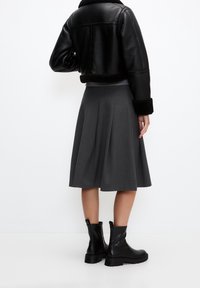 Giacca cropped in pelle nera con dettagli in shearling, abbinata a una gonna a pieghe grigia e stivali neri alla caviglia, vista da dietro.