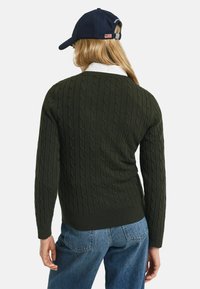 Dunkelgrüner Kabelstrickpullover mit geripptem Saum und Bündchen, getragen über einem weißen Kragenhemd, kombiniert mit blauen Jeans aus Denim und einer marineblauen Mütze.