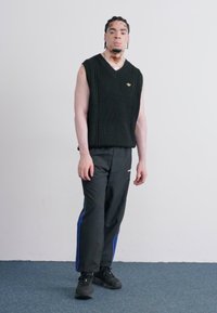 Sort strikket ærmeløs sweater med v-hals, parret med sorte træningsbukser med blå striber i siden, og sorte sneakers. En simpel design.