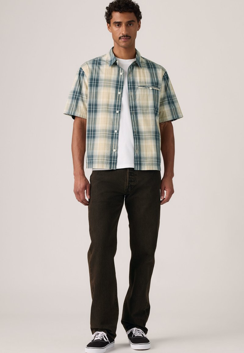 Mand stående iført en kortærmet grøn og beige ternet skjorte over en hvid t-shirt, mørke bukser og sorte sneakers med hvide snørebånd.