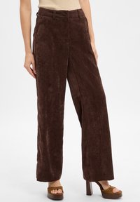 Vrouw in hoge taille, brede bruine ribfluwelen broek met tan open teen platformhakken op een witte achtergrond.