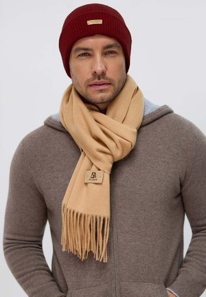 Sciarpa di lana beige con bordi frangiati, etichetta con il marchio, abbinata a un hoodie di maglia marrone e a un berretto a coste bordeaux. Texture morbida.