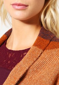 Oranžovo-hnědý pletený svetr s texturovaným vzorem a širokým klopovým límcem. Nositelný přes tmavě bordó krajkový top.