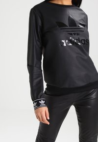 Czarna sportowa bluza z syntetycznego materiału z błyszczącym logo Adidas, okrągłym dekoltem i ściągaczami z białym logo Adidas. Dopasowany krój i faktura.